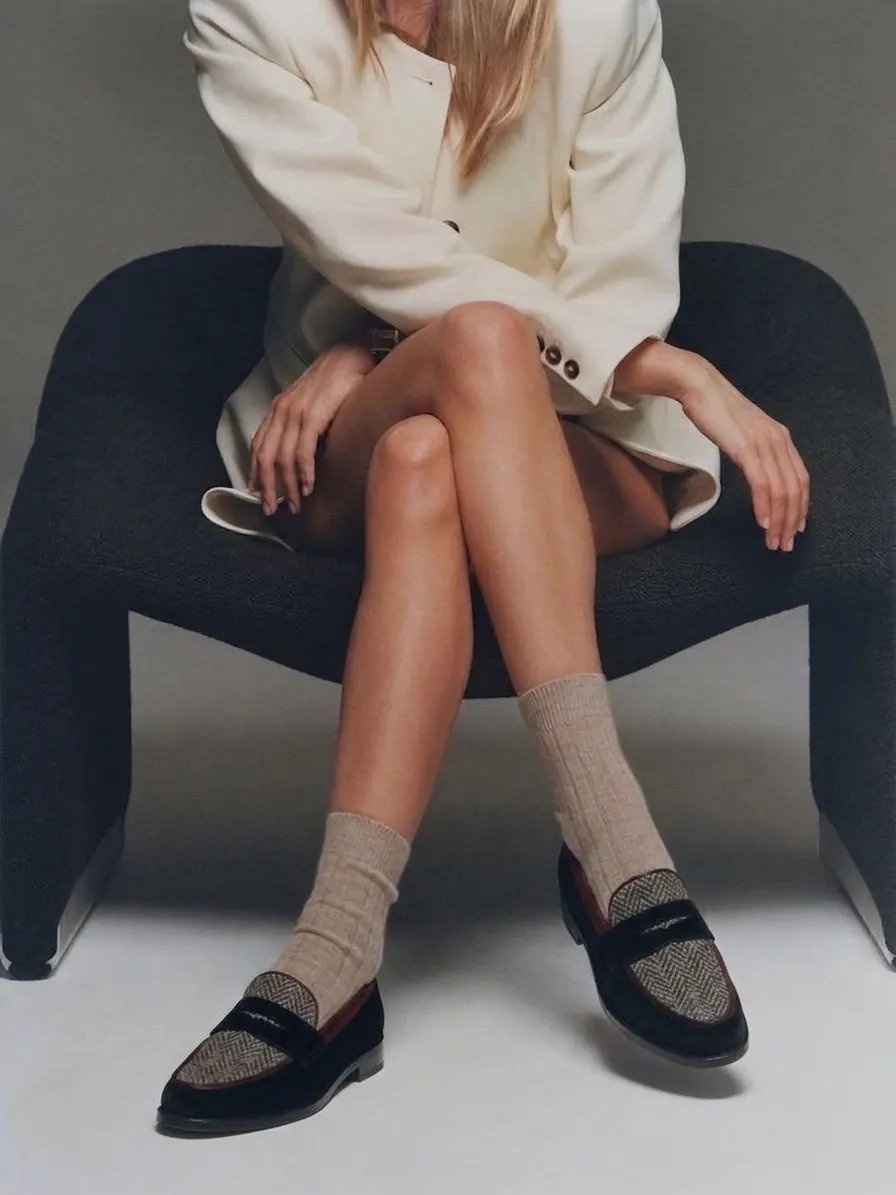Model seated on a dark chair in a cream blazer and LYL herringbone penny loafers